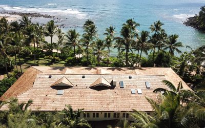 Aerial view of a Hawaii coastal home with solar attic fans installed on a cedar shake roof overlooking the ocean and palm trees.