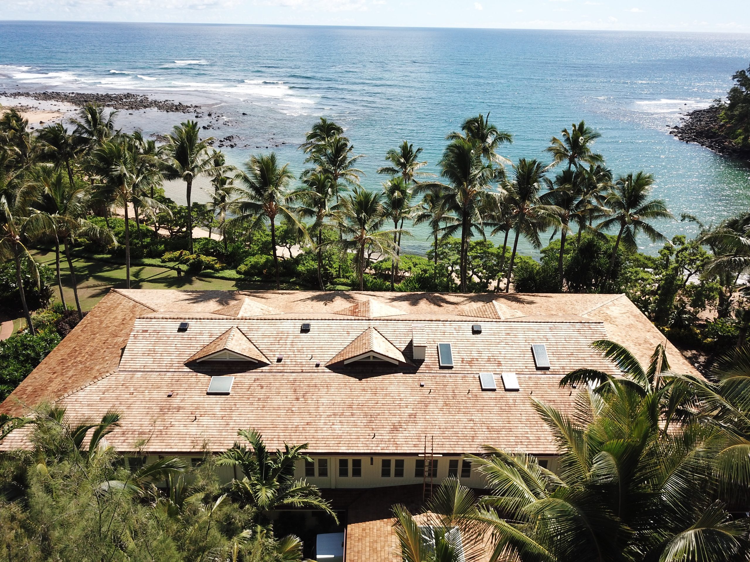 Aerial view of a Hawaii coastal home with solar attic fans installed on a cedar shake roof overlooking the ocean and palm trees.